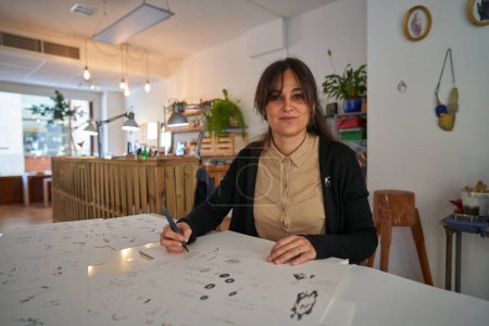 Foto de Mujer con marcador creando bocetos de nuevos accesorios en papeles mientras trabaja en un estudio creativo y mirando a la cámara - Imagen libre de derechos
