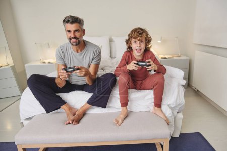 Foto de Longitud completa de hombre descalzo positivo y niño jugando videojuego juntos en el dormitorio - Imagen libre de derechos