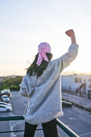 Foto de Vista posterior de la mujer anónima en bufanda rosa de pie en el balcón y levantando el puño por defender contra la enfermedad femenina al atardecer - Imagen libre de derechos