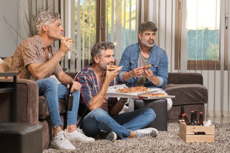 Téléchargez les photos : Jeunes hommes joyeux mangeant de délicieuses pizzas assis dans le salon à la maison et regardant le match de football à la télévision - en image libre de droit