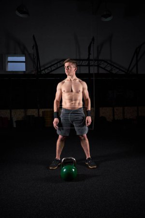 Photo for Full body of confident sportsman with naked torso looking away while standing near kettlebell against dark background - Royalty Free Image