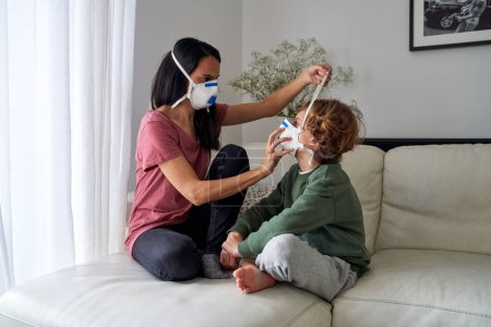 Photo for Full body of focused woman in medical mask putting respirator on face of kid in living room - Royalty Free Image