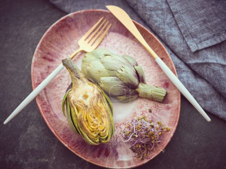 Foto de Desde arriba de corte deliciosas alcachofas servidas en plato con hierbas y cubiertos en la mesa con toalla en cocina ligera - Imagen libre de derechos