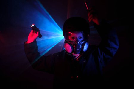 Foto de Niño en máscara de gas con filtros que brillan con linterna mientras está de pie en la oscuridad durante el robo en menta contra la luz de neón azul - Imagen libre de derechos