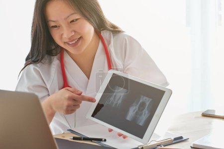 Foto de Cosecha optimista Médico asiático en uniforme médico que apunta a la tableta con rayos X durante la consulta en línea con el paciente en la clínica - Imagen libre de derechos