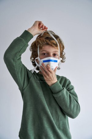 Foto de Niño en suéter verde que se pone mascarilla manteniendo medidas de protección de cuarentena destinadas a la reducción de la propagación de la infección por coronavirus y mirando a la cámara - Imagen libre de derechos