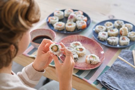 Foto de Alto ángulo de cultivo niño anónimo sentado en la mesa con sushi delicioso y rollos en la mesa en la sala de luz - Imagen libre de derechos