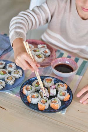 Photo for From above of unrecognizable hungry kid eating sushi rolls with soy sauce using wooden chopsticks in dining room at home - Royalty Free Image