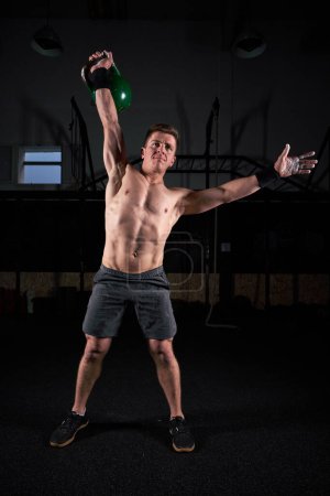 Photo for Full body of fit athlete with naked torso raising am wit heavy kettlebell above head during functional workout in dark gym - Royalty Free Image