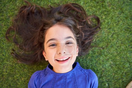 Photo for Top view of happy child with brown eyes resting on green lawn and looking at camera on weekend summer day - Royalty Free Image