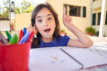 Téléchargez les photos : Adorable enfant de l'école primaire assis à table avec un copybook et des crayons et regardant la caméra avec une idée intelligente dans la tête - en image libre de droit