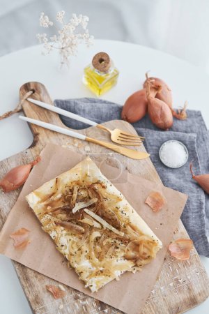 Foto de Desde arriba de hojaldre de cebolla casero tarta servida sobre tabla de cortar de madera con utensilios y cáscara en cocina ligera - Imagen libre de derechos