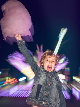 Téléchargez les photos : Garçon incroyable avec bonbons en coton doux regardant la caméra tout en se tenant près de l'attraction lumineuse dans le parc d'attractions en soirée - en image libre de droit
