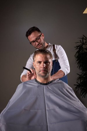 Foto de Peluquero profesional masculino con afeitadora cuello de hombre serio sentado en silla sobre fondo gris en la peluquería moderna luz - Imagen libre de derechos