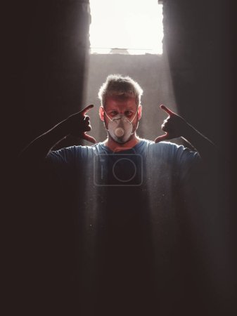 Photo for Adult blond haired male surfboard maker in respirator mask showing Hawaiian Shaka gesture while standing near window illuminated with sunbeam in dark room and looking at camera - Royalty Free Image