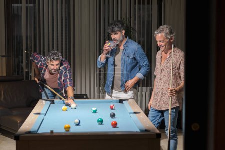 Photo for Positive mature male friends in casual outfits standing near pool table and playing billiards while drinking cocktails in modern room with leather armchair - Royalty Free Image