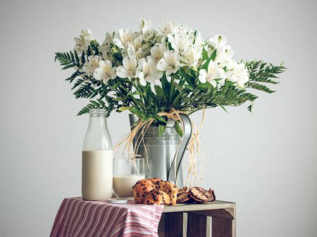 Foto de Bodegón de flores blancas con delicados pétalos en regadera metálica colocado cerca de botella de vidrio de leche y magdalenas en el estudio - Imagen libre de derechos