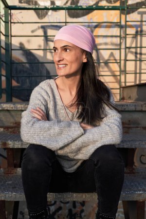 Téléchargez les photos : Heureuse femme hispanique adulte en foulard rose pour prêter attention au cancer du sein féminin assis avec les bras croisés sur un escalier métallique minable et regardant ailleurs - en image libre de droit