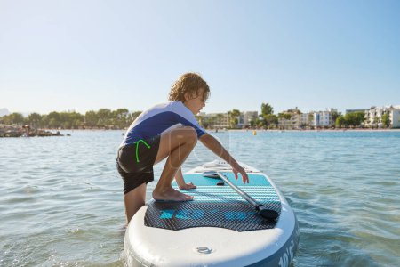 Photo for Side view of boy in activewear climbing on paddle surfboard in middle of ocean with beach in background against cloudless sky - Royalty Free Image