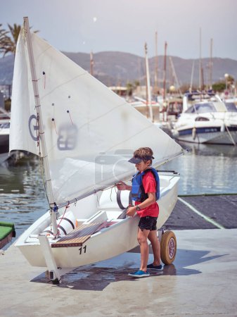 Photo for Full body side view of male child in protective vest preparing sailboat on harbor near lake during summer vacation - Royalty Free Image