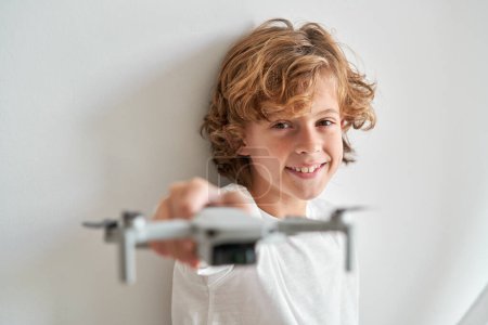Photo for Happy child with curly hair looking at camera while stretching hand with blurred toy drone near white wall - Royalty Free Image