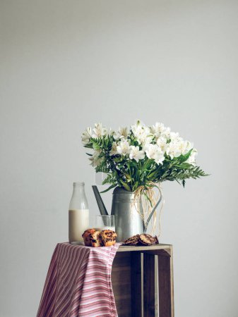 Photo for Bunch of flowers with white blossoms in metal watering can placed on wooden crate with bottle of milk and tasty pastry - Royalty Free Image