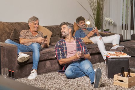 Téléchargez les photos : Souriant amis masculins utilisant des smartphones assis dans le salon confortable pendant la fête de week-end - en image libre de droit