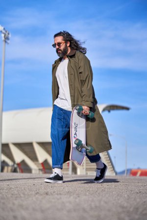 Photo for From below of full body stylish mature bearded male in sunglasses wearing trendy raincoat and jeans and sneakers walking on road with longboard in sunny day - Royalty Free Image
