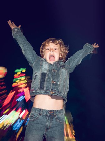 Photo for Joyful boy with raised arms looking at camera with opened mouth while standing near attraction in amusement park in evening time - Royalty Free Image