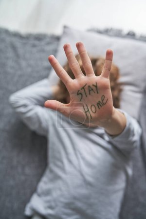 Photo for From above of anonymous child showing hand with Stay Home writing while lying on blanket and pillow during pandemic at home - Royalty Free Image
