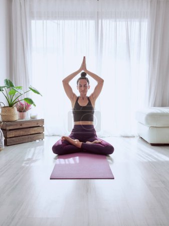 Téléchargez les photos : Fit femelle en vêtements de sport méditant dans la pose Lotus avec les bras levés tout en pratiquant le yoga à la maison - en image libre de droit