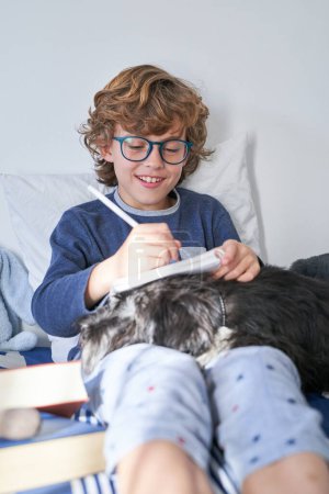 Téléchargez les photos : Garçon positif dans les lunettes assis avec chien moelleux et prendre des notes dans le carnet tout en faisant une tâche et étudier dans la chambre à coucher à la maison - en image libre de droit