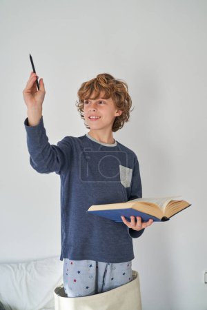 Photo for Delightful boy in casual clothes looking away while holding open book and raising arm with pencil in light room - Royalty Free Image