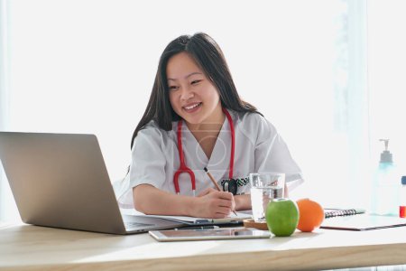 Téléchargez les photos : Souriant asiatique femme médecin en uniforme écriture sur presse-papiers tout en surfant netbook à table avec tablette dans la clinique de lumière moderne - en image libre de droit