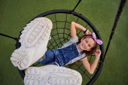 Photo for Top view of joyful kid in overall looking at camera while lying on web swing with raised legs on playground in summer - Royalty Free Image