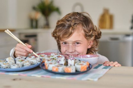 Foto de Niño juguetón con el pelo rizado sonriendo y mirando a la cámara mientras come rollos de sushi con palillos y se esconde detrás de los platos en la cocina en casa - Imagen libre de derechos