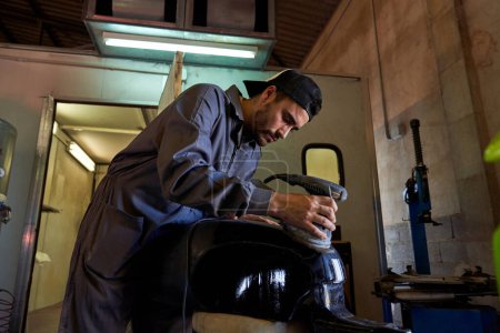 Foto de Maestro masculino enfocado en tanque de motocicleta de rectificado uniforme con máquina de pulido mientras trabaja en taller profesional en luz tenue - Imagen libre de derechos