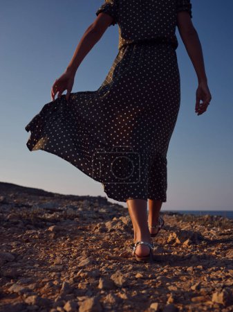 Téléchargez les photos : D'en bas de femme méconnaissable en robe à pois élégant marchant le long de la côte rocheuse près de la mer au coucher du soleil - en image libre de droit