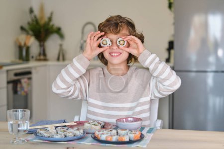 Photo for Positive blond boy having fun with rolls while sitting at table with plate of tasty sushi in light kitchen - Royalty Free Image