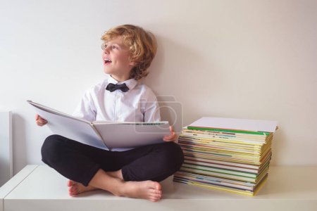 Photo for Full body of cheerful boy in formal wear looking into distance with smile while reading book on white background near heap of textbooks - Royalty Free Image