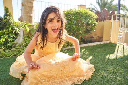 Photo for Ground level of mischievous preteen kid in elegant yellow dress screaming and looking at camera while sitting on grass in garden - Royalty Free Image