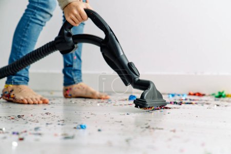 Photo for Crop anonymous barefoot boy in blue jeans using vacuum cleaner for collecting colorful confetti from floor after party - Royalty Free Image