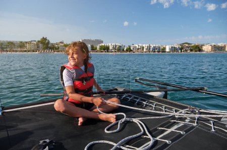 Photo for Full length of content barefooted preteen kid with blond hair in life vest smiling and looking away while sitting on boat and pulling rope during summer vacation - Royalty Free Image