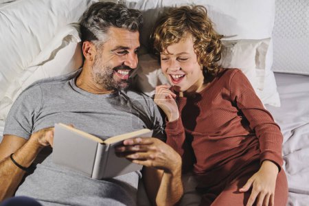 From above of happy father and son in pajamas having fun while reading interesting book and lying on soft bed together at home