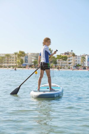 Foto de Vista trasera del niño parado en el paddleboard y remando en el océano ondulado mientras mira la cámara contra el cielo despejado durante las vacaciones de verano - Imagen libre de derechos