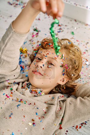 Photo for High angle of curly haired boy lying on floor with hand behind head and playing with colorful confetti while looking at camera - Royalty Free Image