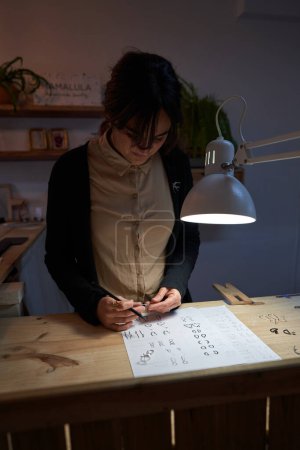 Foto de Hembra orfebre concentrada de pie con lápiz a la mesa con dibujos dibujados iluminados con lámpara y mirando el anillo en las manos - Imagen libre de derechos