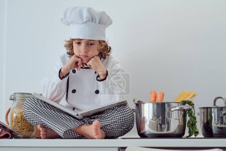 Téléchargez les photos : De dessous de préadolescent réfléchi garçon en costume de chef et casquette assis avec les jambes croisées et ouvert livre de cuisine près de la casserole avec des carottes et des spaghettis - en image libre de droit