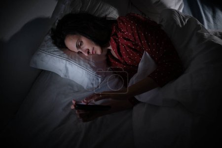Photo for From above of serious Hispanic female in sleepwear text messaging on cellphone while lying on bed late at night time - Royalty Free Image