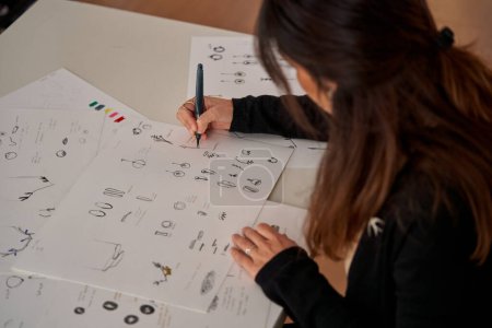 Foto de Alto ángulo de la cosecha anónima diseñadora femenina dibujo bocetos para accesorios hechos a mano en papel en el taller - Imagen libre de derechos